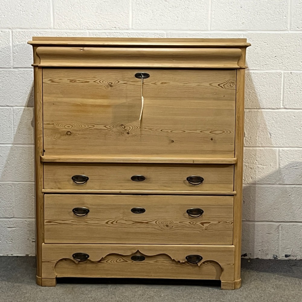 A tall antique stripped pine secretaire bureau featuring a upper fall-front writing flap above three long drawers with brass handles and a carved lower apron.