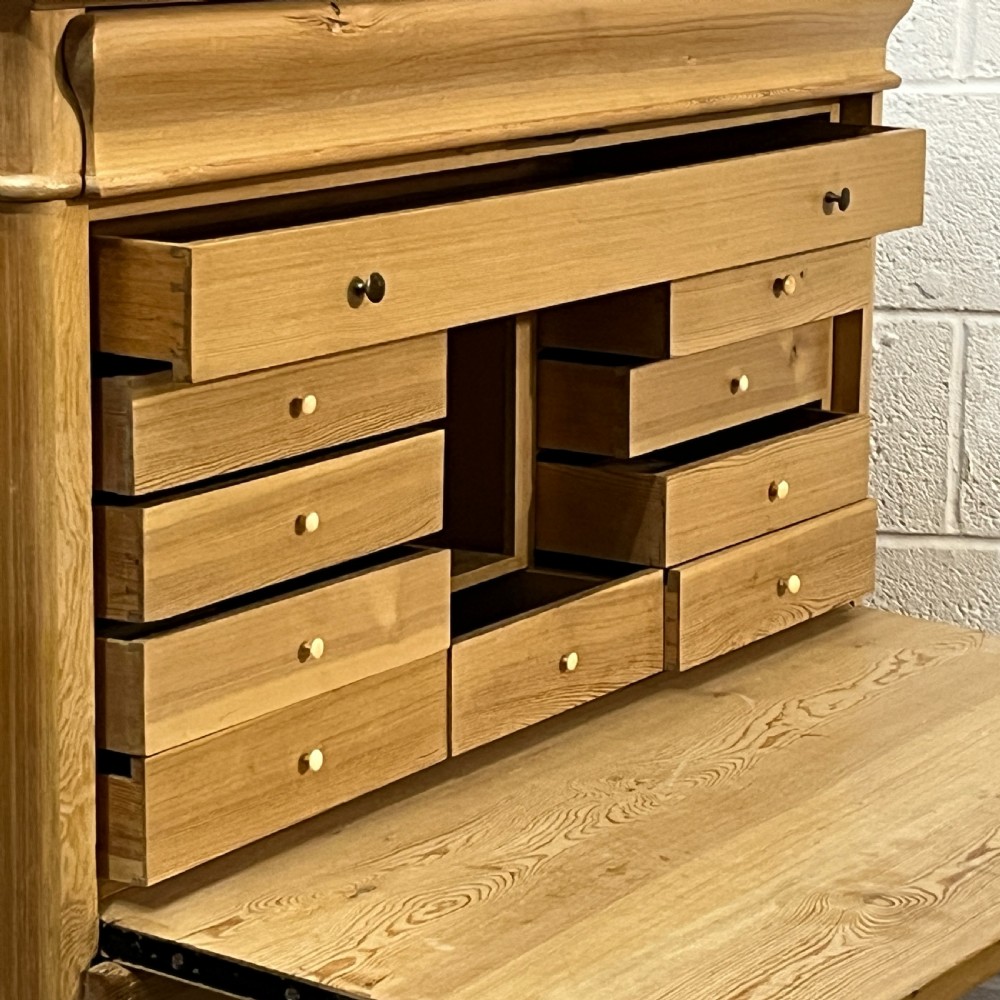 A tall antique stripped pine secretaire bureau featuring a upper fall-front writing flap above three long drawers with brass handles and a carved lower apron. View IX