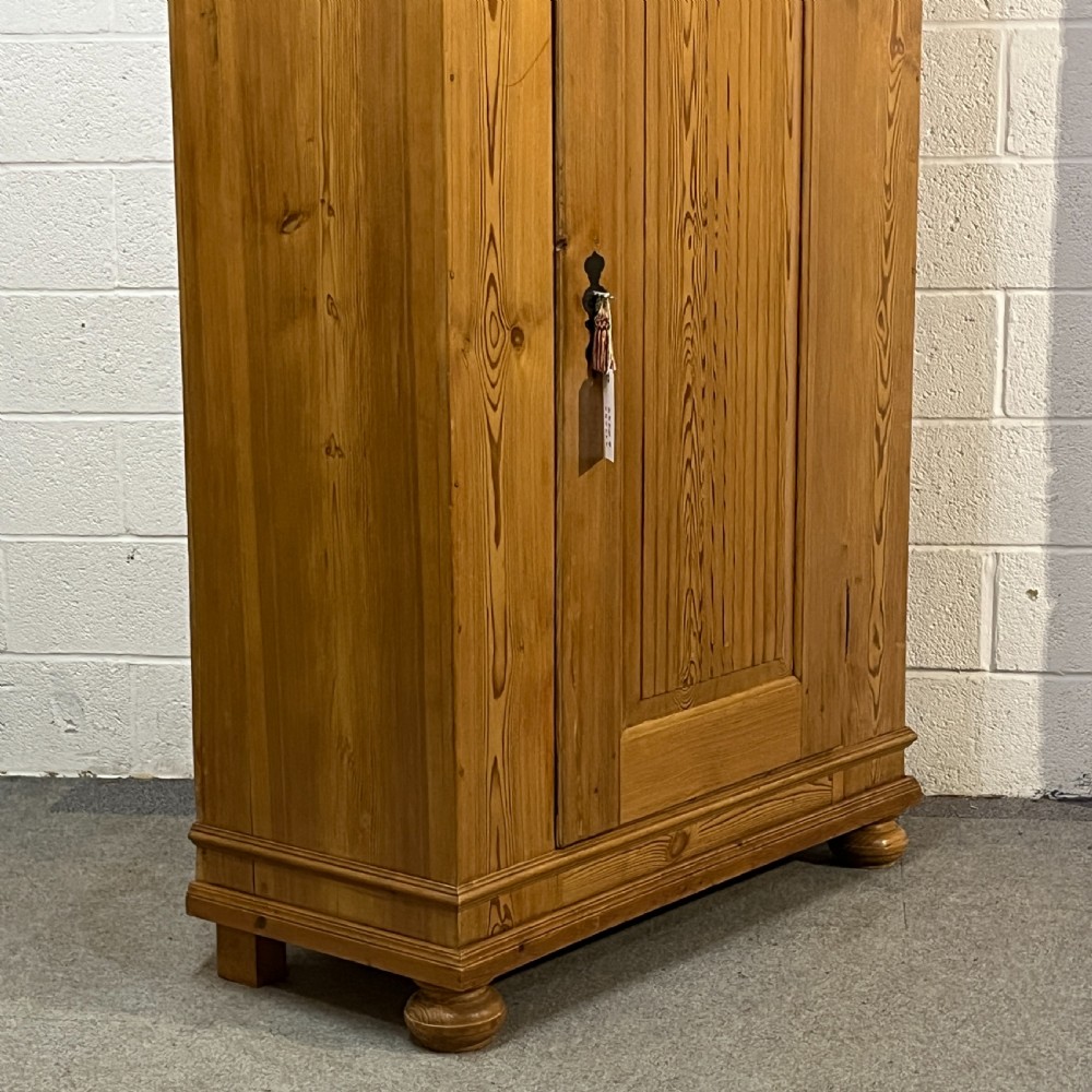 A tall antique waxed pine storage cupboard featuring a single panelled door, an iron keyhole escutcheon, a moulded top cornice, and bun feet. View V