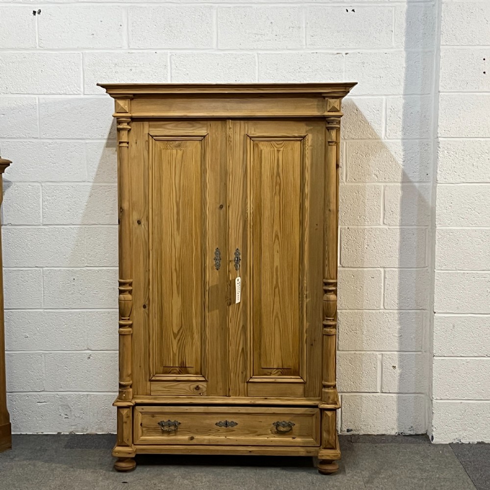 A freestanding double-door antique pine wardrobe featuring turned front columns, panelled doors, key escutcheons, and a single bottom drawer.