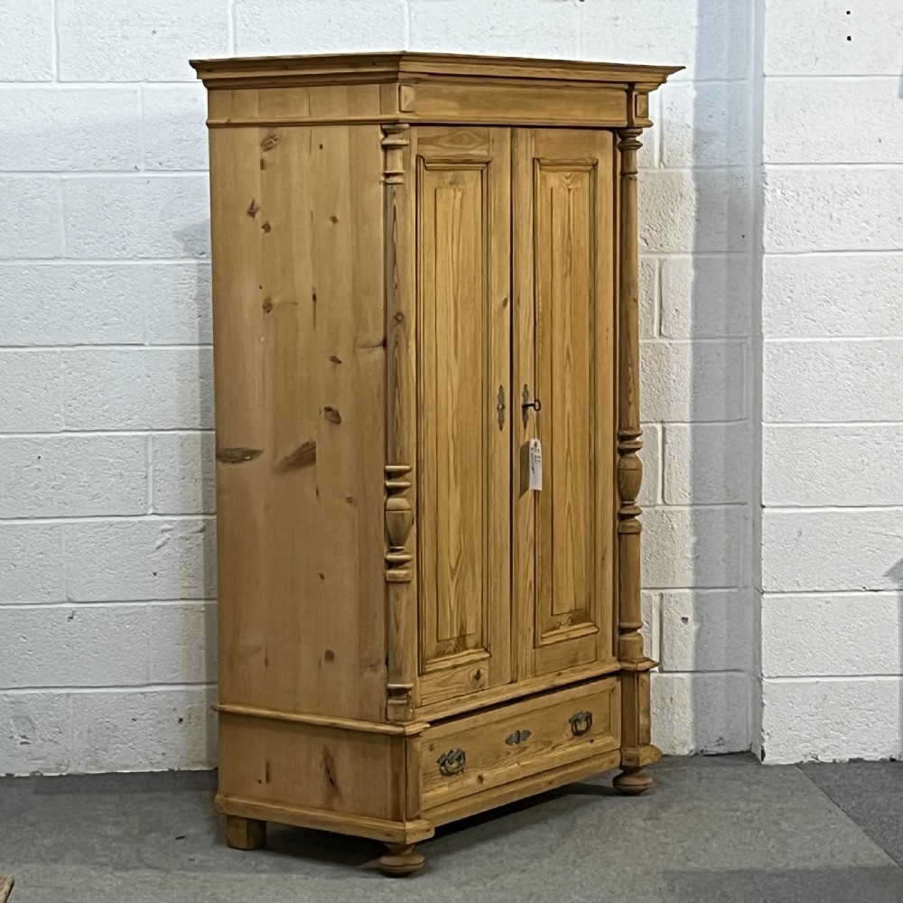 A freestanding double-door antique pine wardrobe featuring turned front columns, panelled doors, key escutcheons, and a single bottom drawer. View III