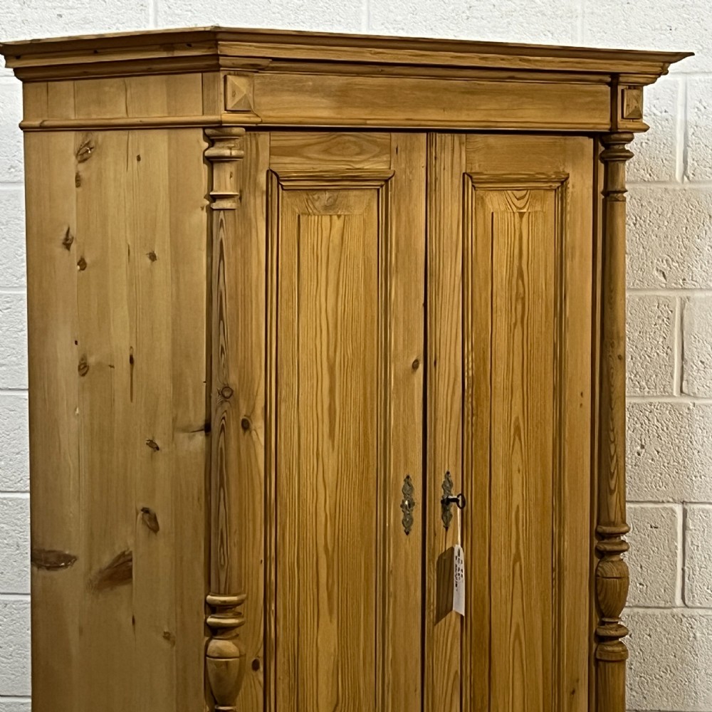 A freestanding double-door antique pine wardrobe featuring turned front columns, panelled doors, key escutcheons, and a single bottom drawer. View V
