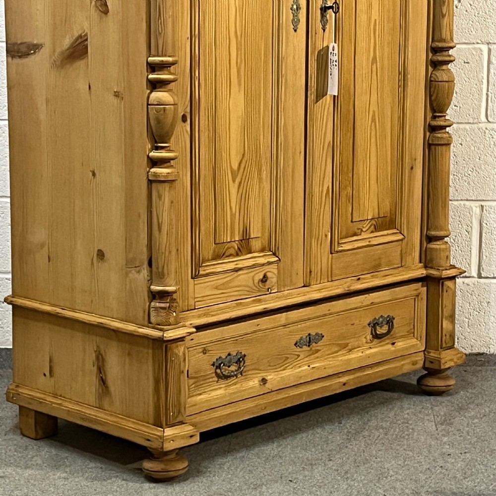A freestanding double-door antique pine wardrobe featuring turned front columns, panelled doors, key escutcheons, and a single bottom drawer. View VI