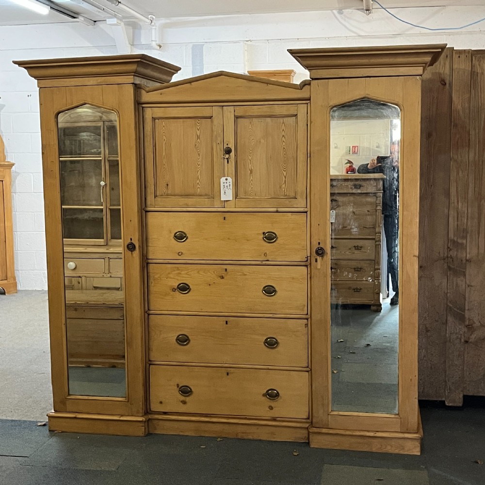 A large Victorian pine combination wardrobe featuring a central chest of drawers and upper cupboard, flanked by two full-length mirrored side doors with arched tops.