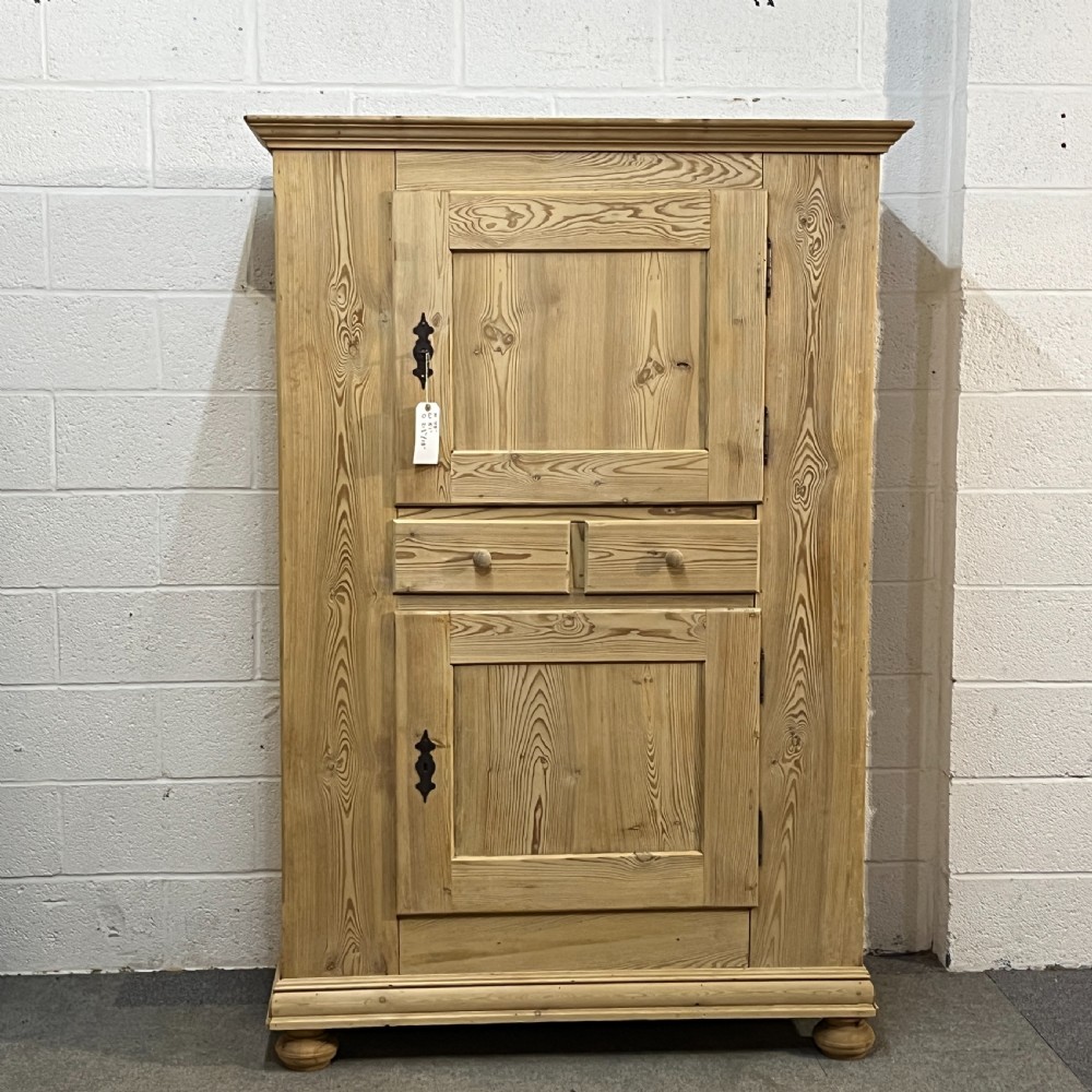 A tall stripped antique pine storage cupboard featuring two fielded panel doors, two central drawers, black iron escutcheons, and turned bun feet.