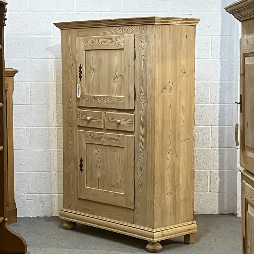 A tall stripped antique pine storage cupboard featuring two fielded panel doors, two central drawers, black iron escutcheons, and turned bun feet. View II