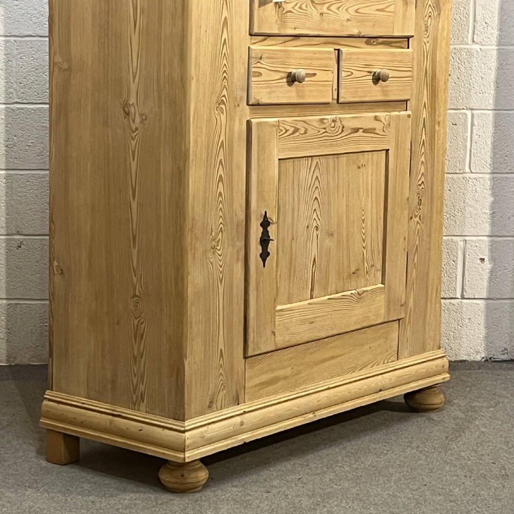 A tall stripped antique pine storage cupboard featuring two fielded panel doors, two central drawers, black iron escutcheons, and turned bun feet. View V