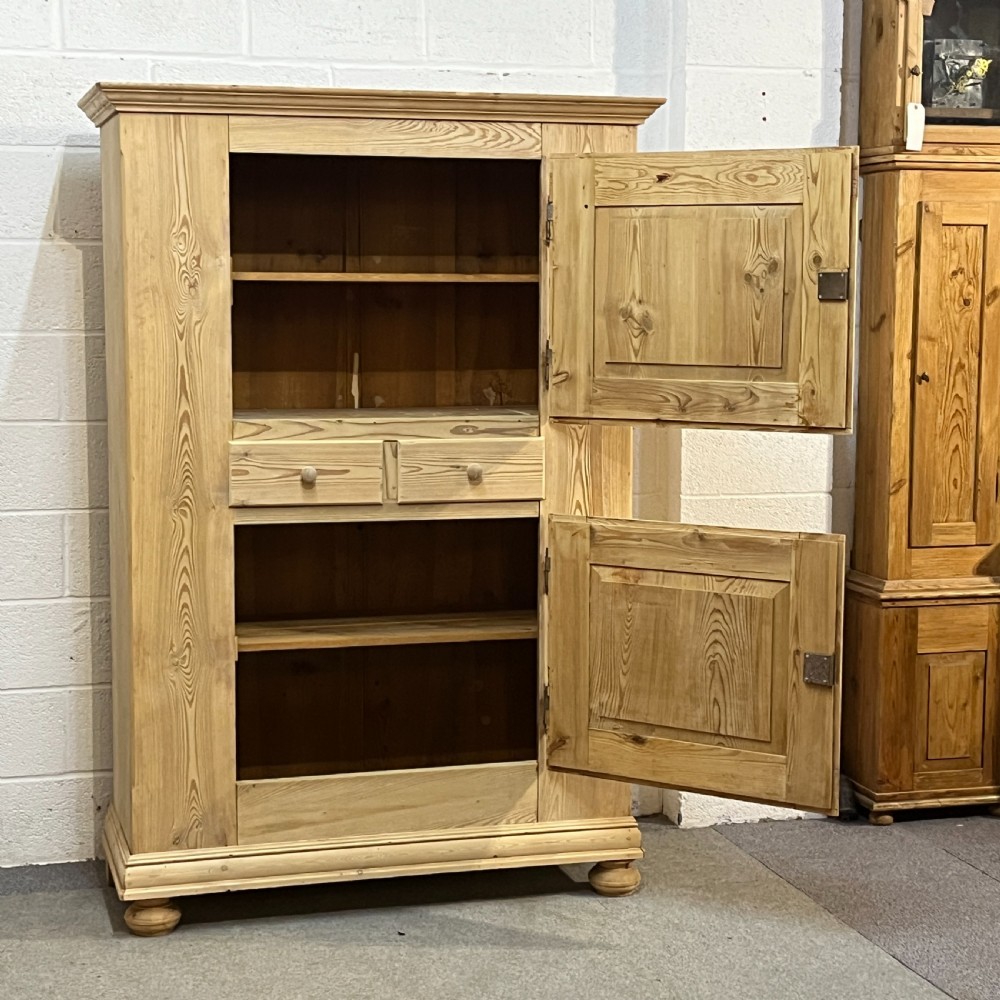 A tall stripped antique pine storage cupboard featuring two fielded panel doors, two central drawers, black iron escutcheons, and turned bun feet. View VI