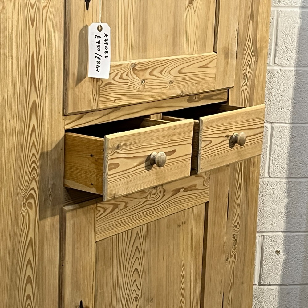 A tall stripped antique pine storage cupboard featuring two fielded panel doors, two central drawers, black iron escutcheons, and turned bun feet. View VIII
