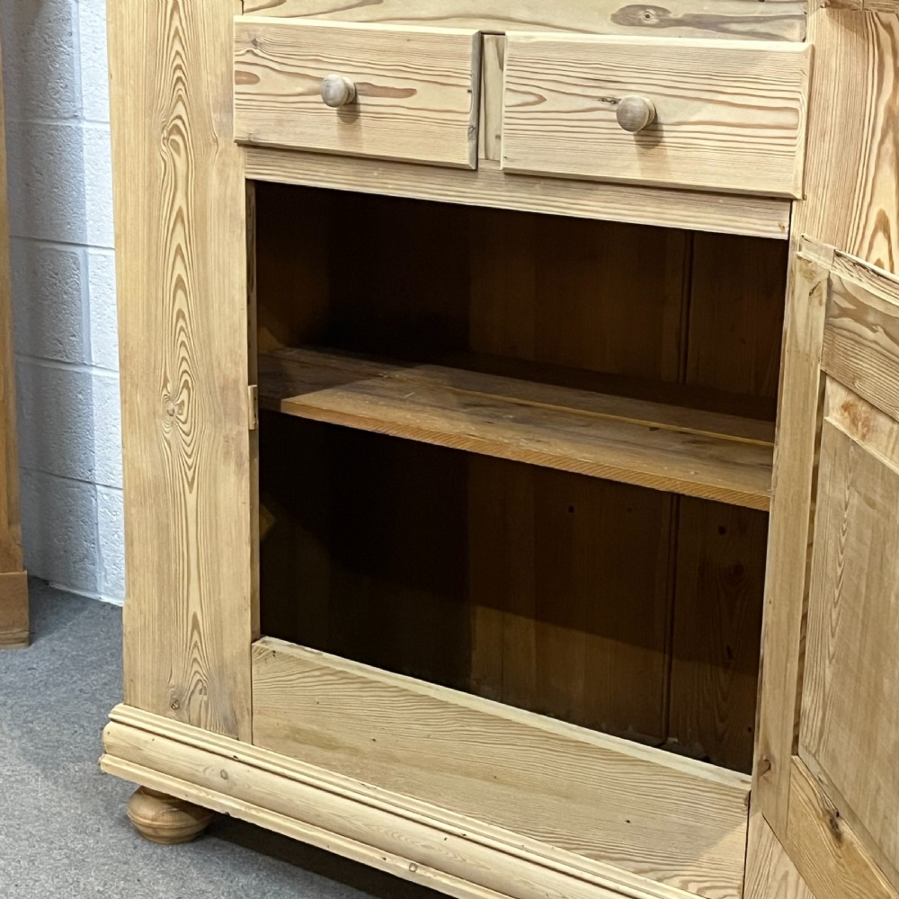 A tall stripped antique pine storage cupboard featuring two fielded panel doors, two central drawers, black iron escutcheons, and turned bun feet. View IX