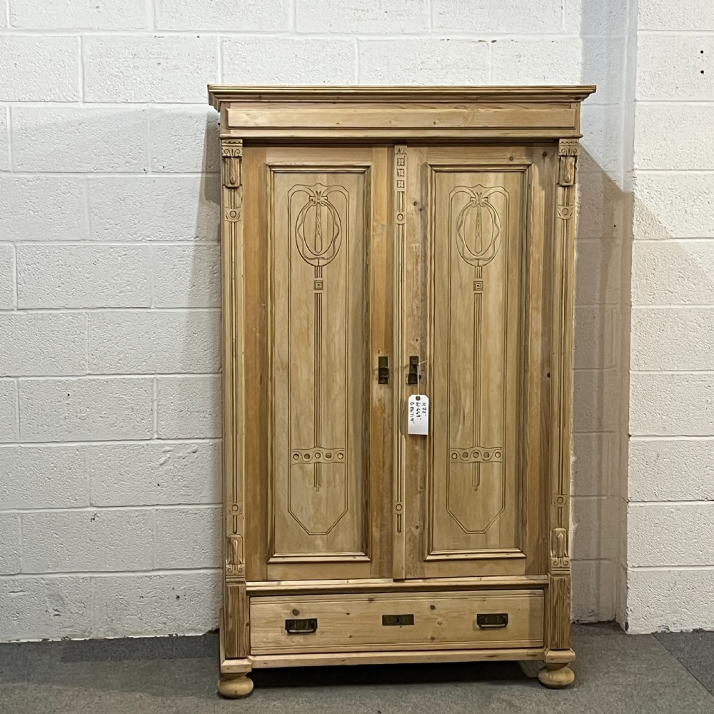 A stripped pine wardrobe featuring carved panel doors with Art Nouveau motifs, a lower drawer with brass handles, and turned bun feet.