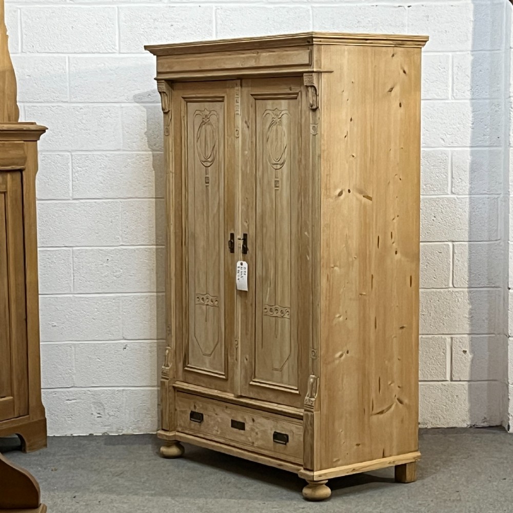 A stripped pine wardrobe featuring carved panel doors with Art Nouveau motifs, a lower drawer with brass handles, and turned bun feet. View II