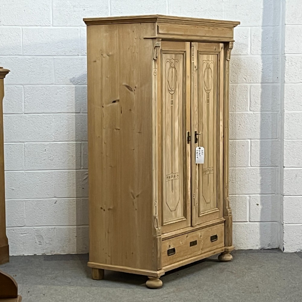 A stripped pine wardrobe featuring carved panel doors with Art Nouveau motifs, a lower drawer with brass handles, and turned bun feet. View III