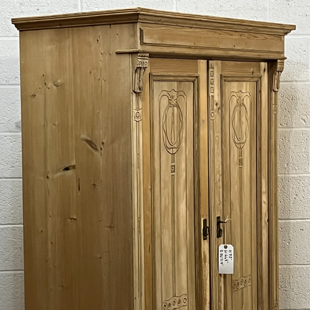 A stripped pine wardrobe featuring carved panel doors with Art Nouveau motifs, a lower drawer with brass handles, and turned bun feet. View V