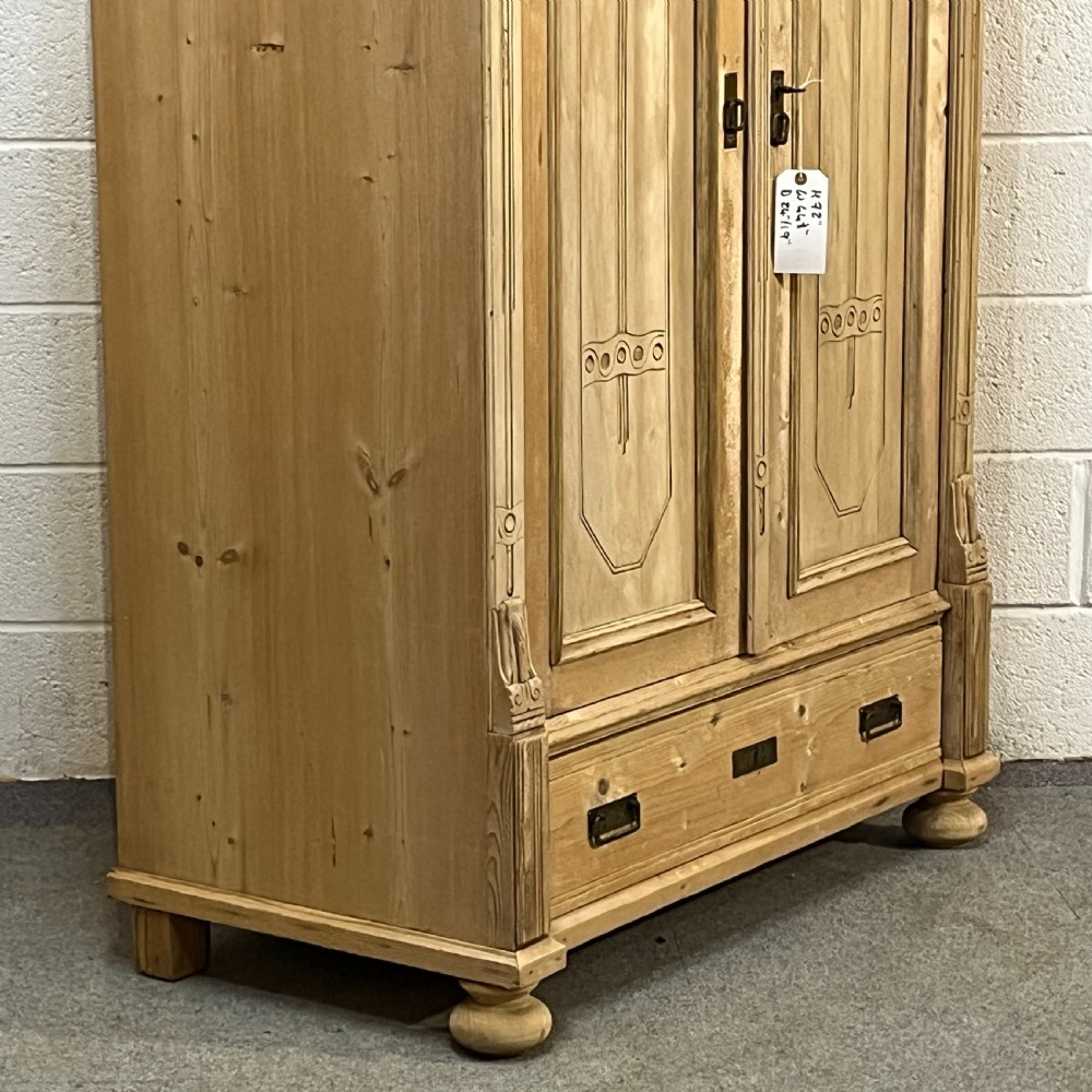 A stripped pine wardrobe featuring carved panel doors with Art Nouveau motifs, a lower drawer with brass handles, and turned bun feet. View VI