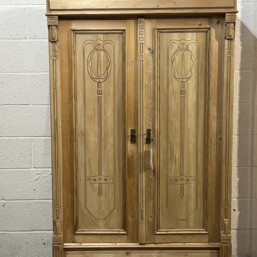 A stripped pine wardrobe featuring carved panel doors with Art Nouveau motifs, a lower drawer with brass handles, and turned bun feet. View VII