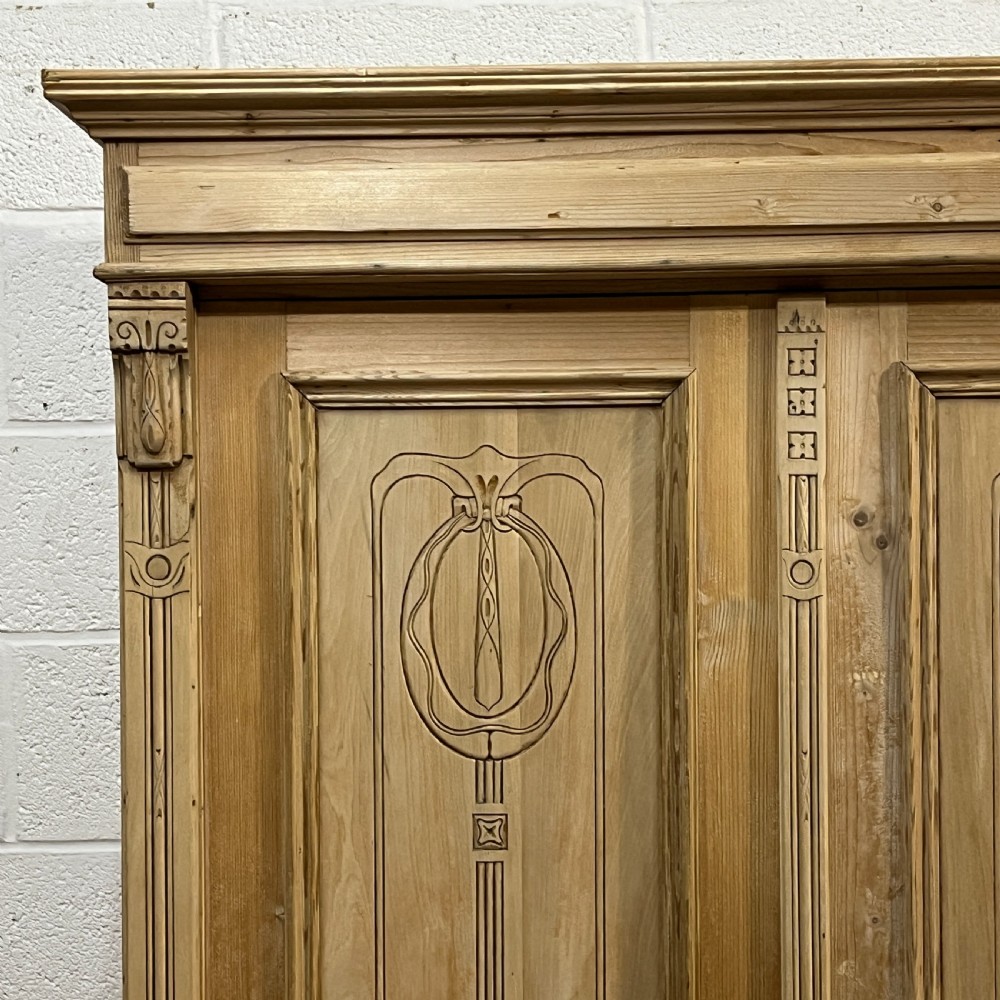 A stripped pine wardrobe featuring carved panel doors with Art Nouveau motifs, a lower drawer with brass handles, and turned bun feet. View IX