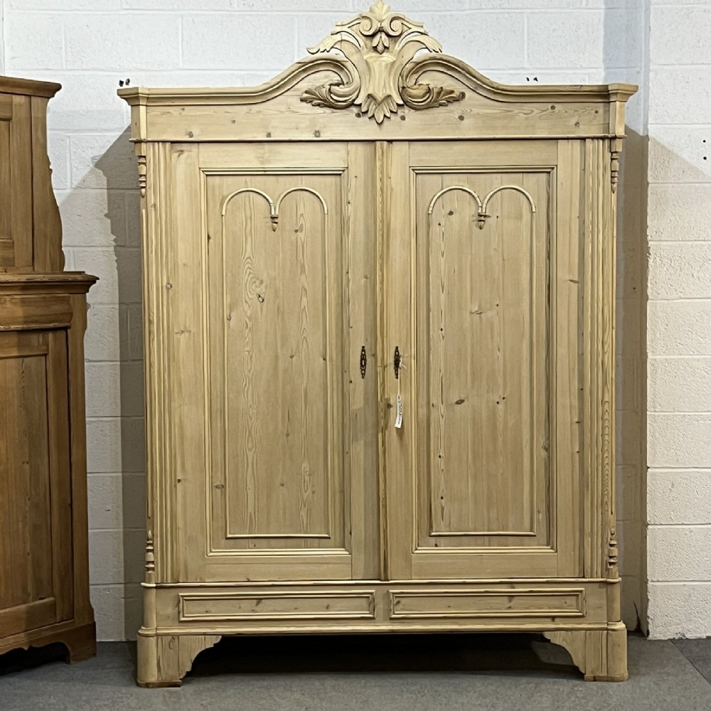 A large late nineteenth-century natural pine double wardrobe featuring a carved scroll pediment, arched door mouldings, and a flat plinth base.