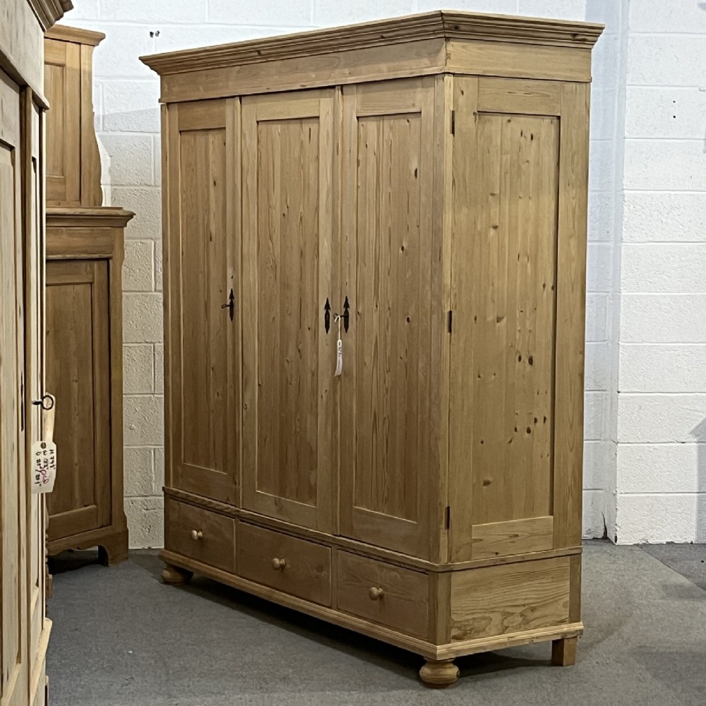A large natural pine triple wardrobe featuring three vertical panelled doors above three aligned base drawers with turned wooden knobs. View VI