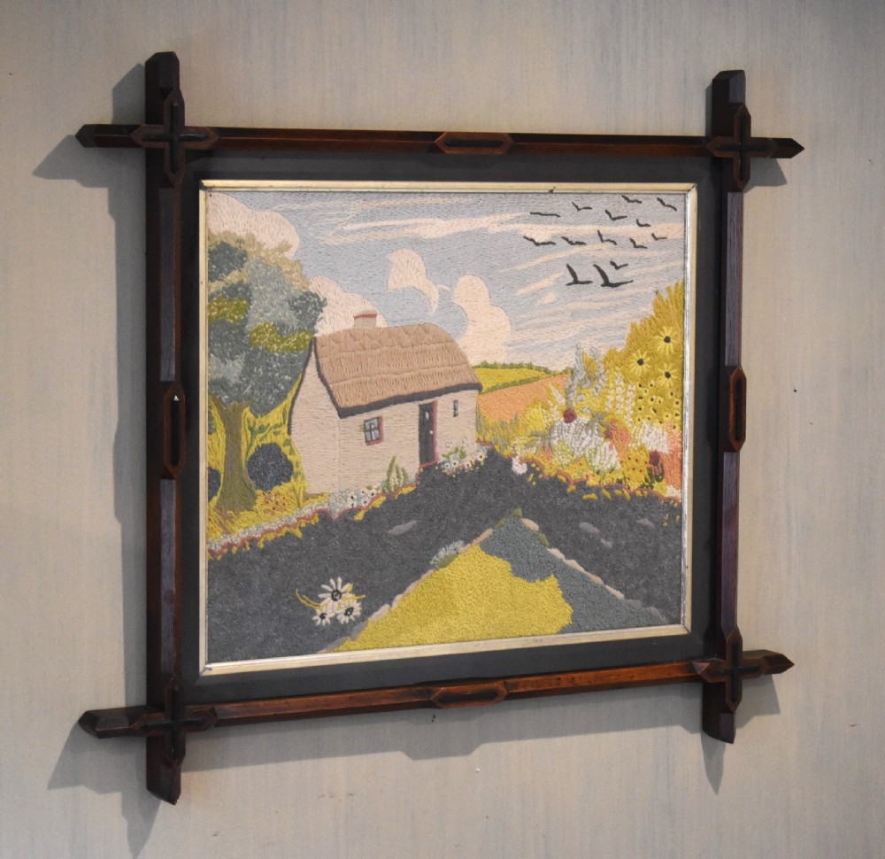 An Arts and Crafts needlework picture depicting a thatched cottage landscape framed in a dark stained wood frame with crossed corner joints.