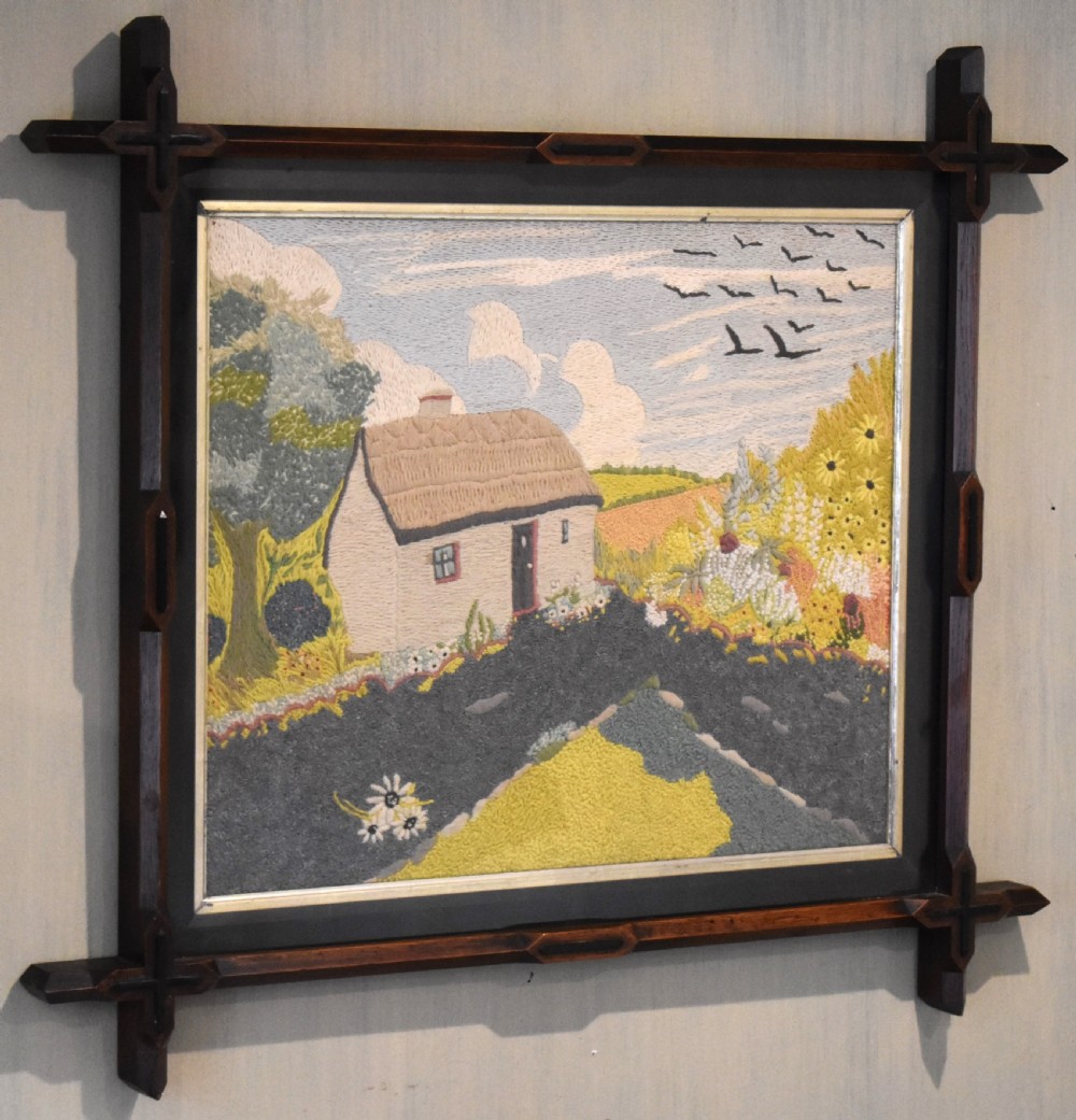 An Arts and Crafts needlework picture depicting a thatched cottage landscape framed in a dark stained wood frame with crossed corner joints. View II