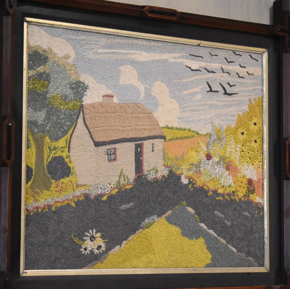 An Arts and Crafts needlework picture depicting a thatched cottage landscape framed in a dark stained wood frame with crossed corner joints. View III