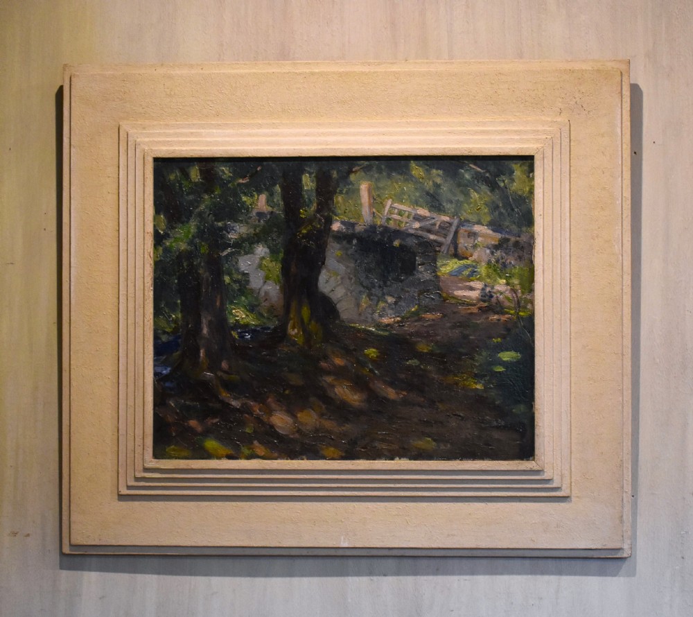 An oil painting depicting a dark woodland scene with trees, a stone bridge, and a wooden gate, housed within a textured off-white stepped frame.