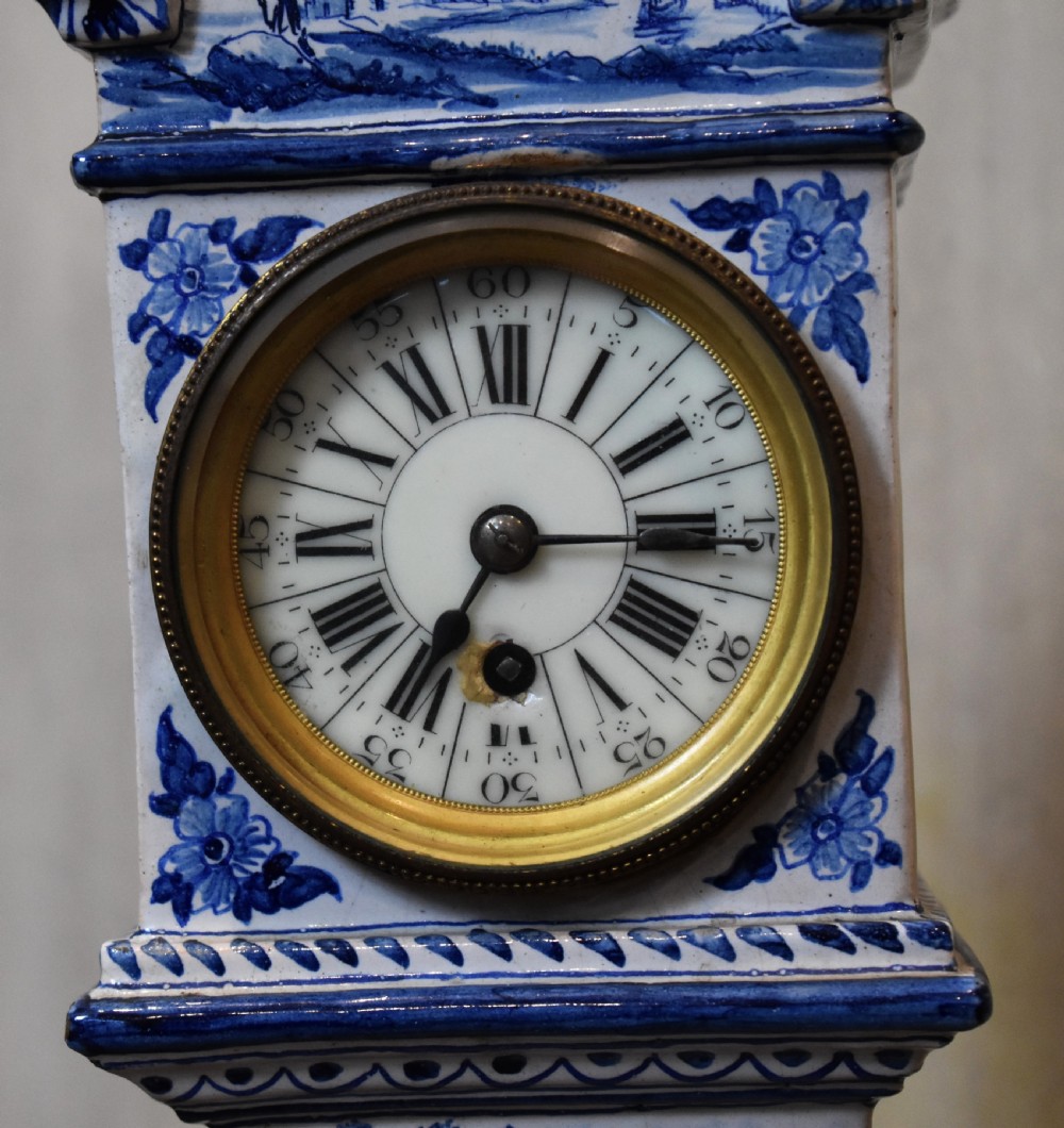 A blue and white tin-glazed ceramic miniature longcase clock featuring hand-painted rural figures, floral motifs, and a white clock face with Roman numerals framed in brass. View IV