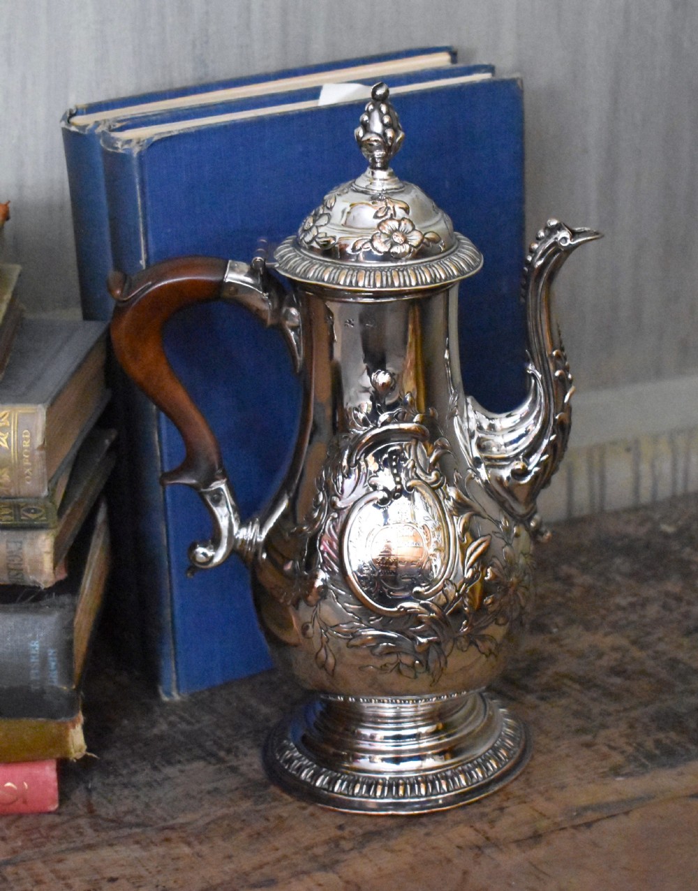 An antique Georgian silver-plated coffee pot featuring repousse floral scrollwork, an engraved cartouche, a carved wooden handle, and a finial lid.