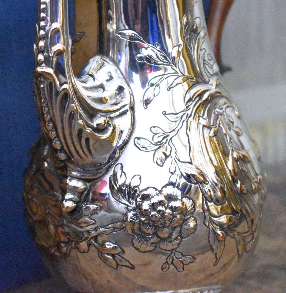 An antique Georgian silver-plated coffee pot featuring repousse floral scrollwork, an engraved cartouche, a carved wooden handle, and a finial lid. View V