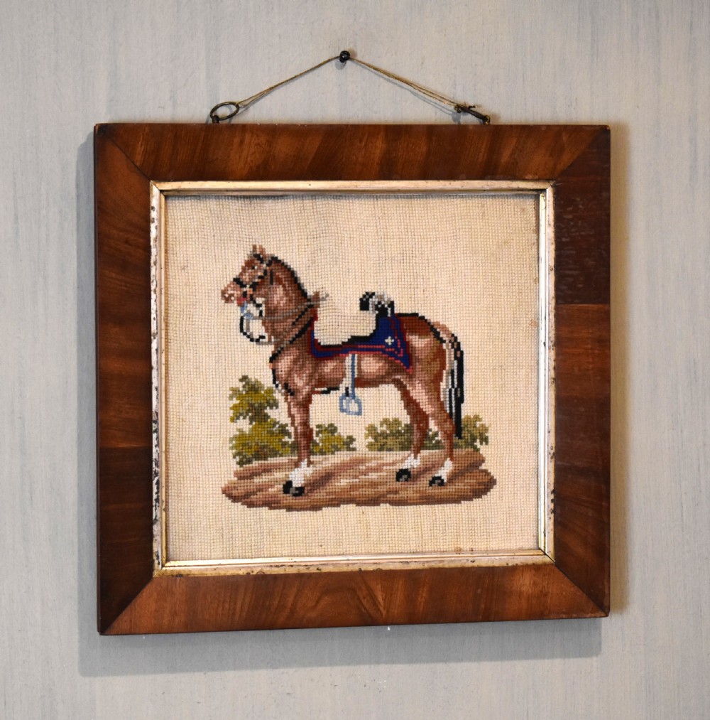 A square framed Victorian needlework picture depicting a brown saddled horse standing in a landscape within a wide wooden frame with a gilded inner slip.