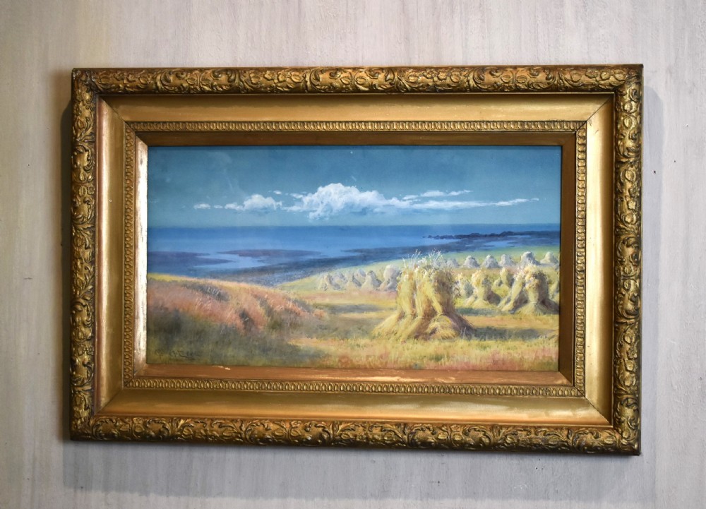 A framed watercolour painting depicting hay stooks in a field overlooking a blue bay under a cloudy sky, presented in a decorative gilt frame.