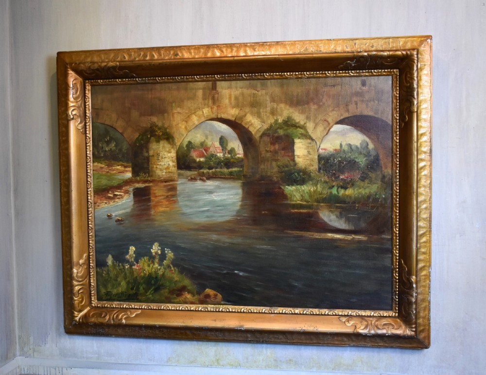 A framed oil painting featuring a large stone arch bridge over a river with reflections and a distant village.