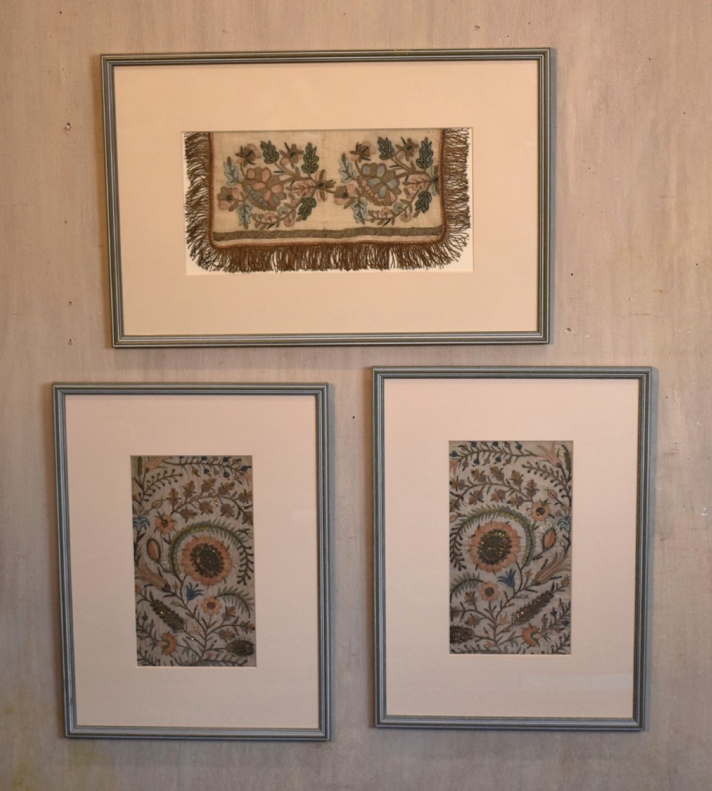 Set of three framed antique needlework panels displaying intricate botanical embroidery, mounted in cream pass-partout and light grey frames.