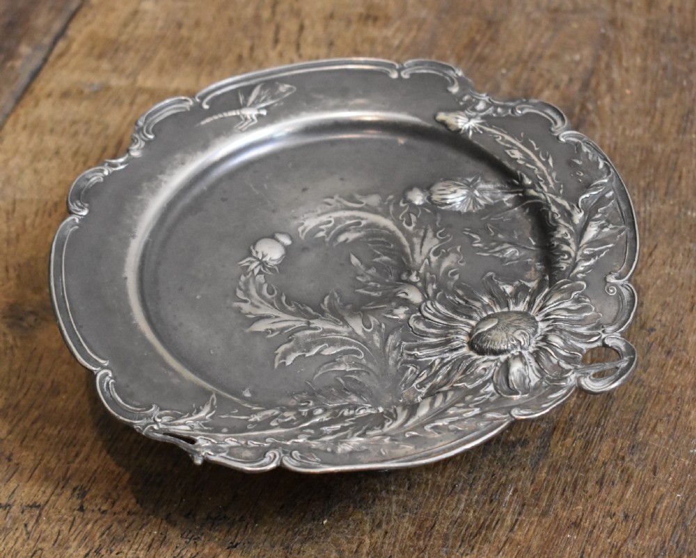 An Art Nouveau Britannia metal tazza with relief carvings of flowers, foliage, and a dragonfly along the scalloped rim.