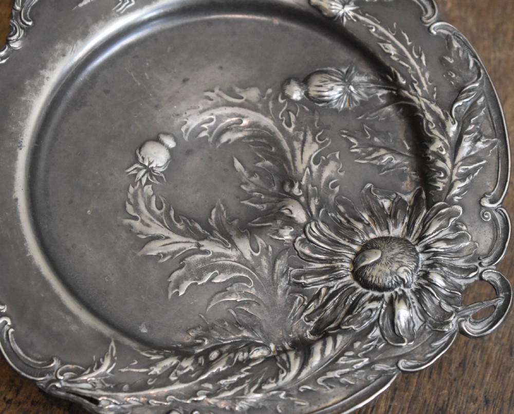 An Art Nouveau Britannia metal tazza with relief carvings of flowers, foliage, and a dragonfly along the scalloped rim. View IV