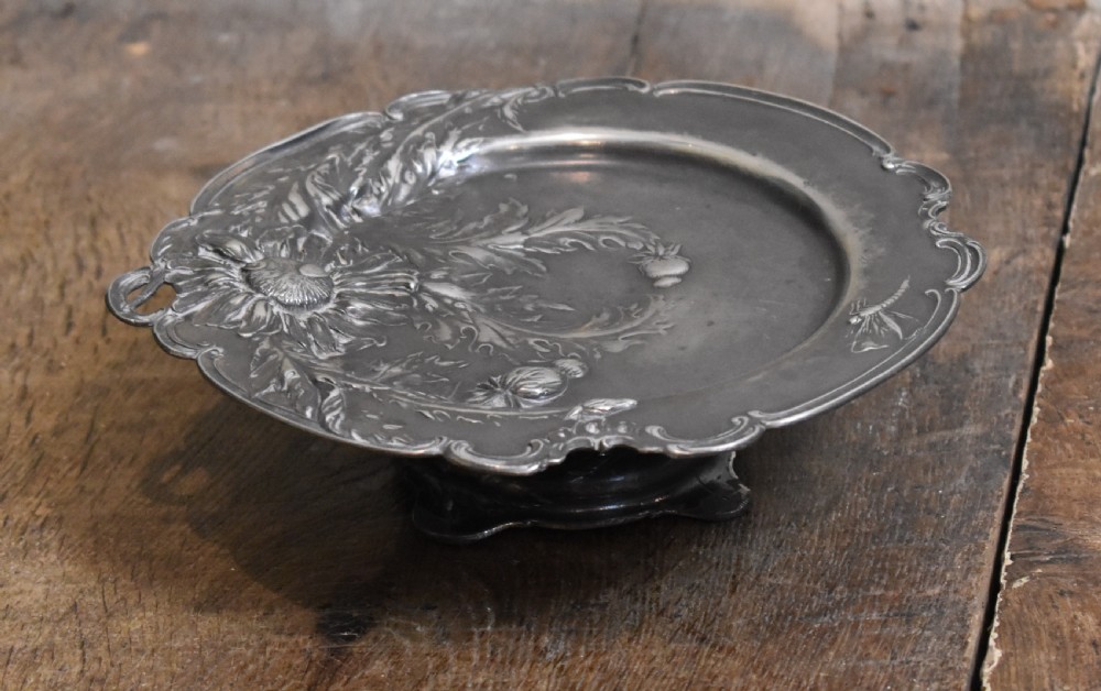 An Art Nouveau Britannia metal tazza with relief carvings of flowers, foliage, and a dragonfly along the scalloped rim. View V
