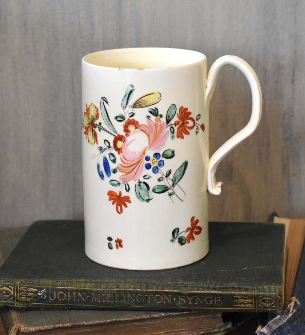 A cylindrical creamware ceramic tankard decorated with hand-painted polychrome floral motifs and a loop handle.