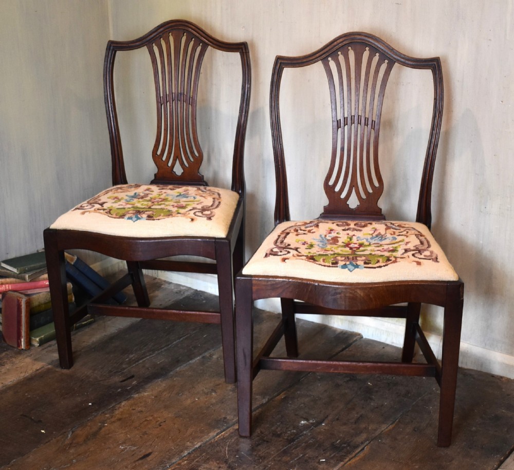 A pair of Georgian mahogany side chairs featuring carved pierced splat backs, serpentine top rails, and embroidered drop-in seats.