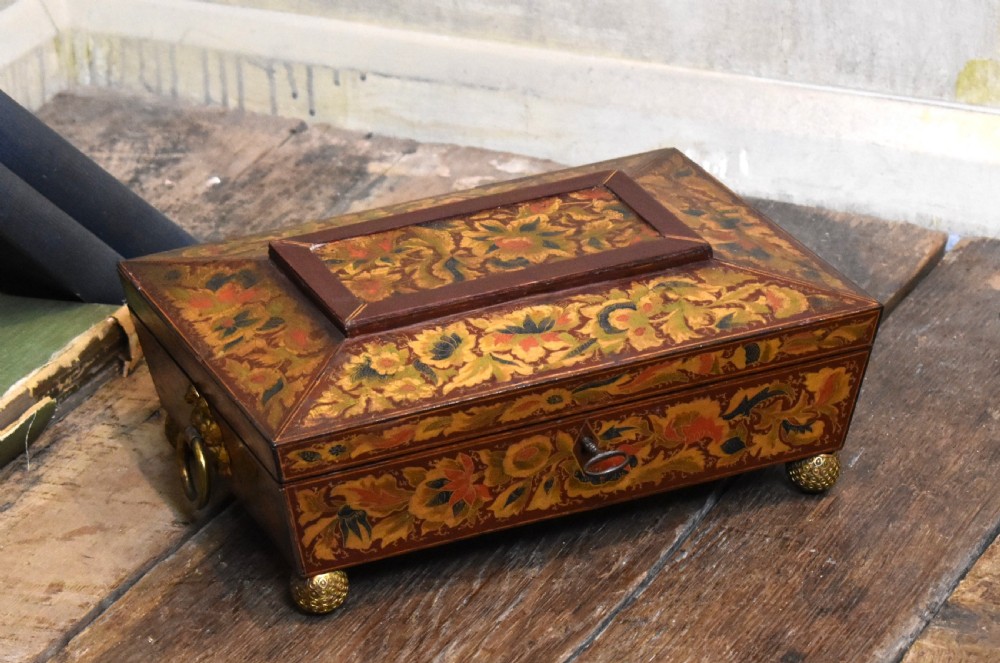 A rectangular Regency period wooden box featuring intricate botanical penwork decoration, brass ring handles, and ball feet.
