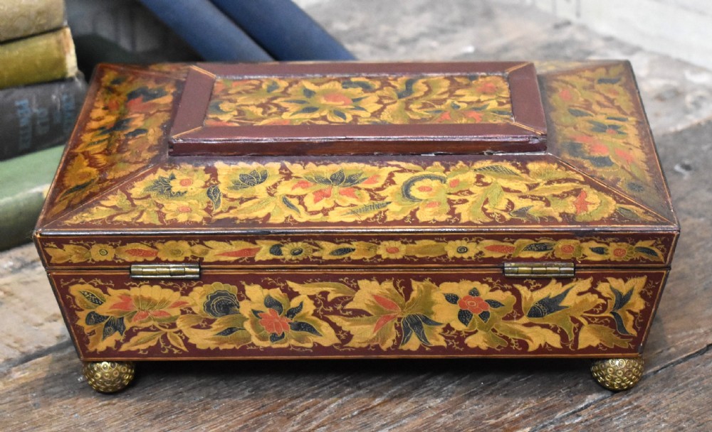 A rectangular Regency period wooden box featuring intricate botanical penwork decoration, brass ring handles, and ball feet. View V