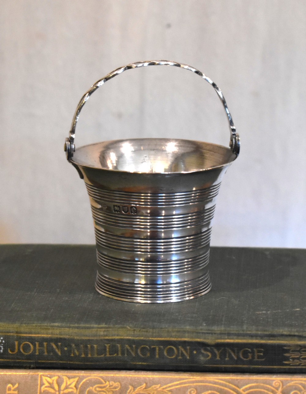 A solid silver miniature bucket pail featuring horizontal reeded detailing, visible hallmarks on the exterior, and a twisted swing handle.