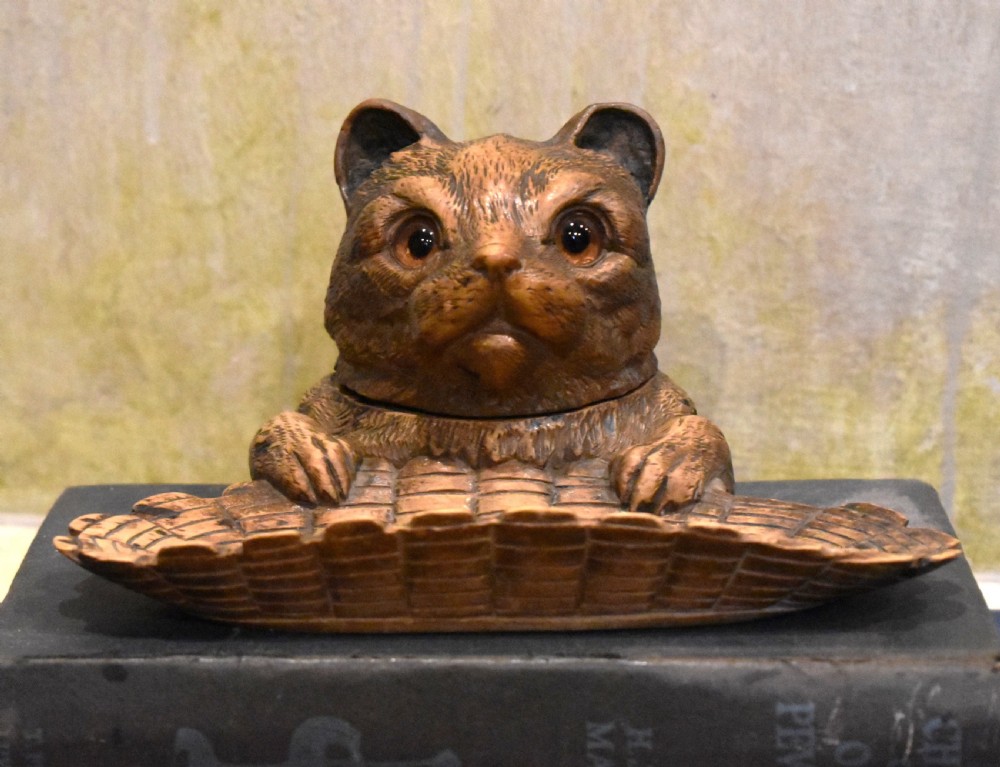 A carved wooden Black Forest inkwell featuring the detailed head of a cat with glass eyes positioned behind a basketweave pen tray. View II