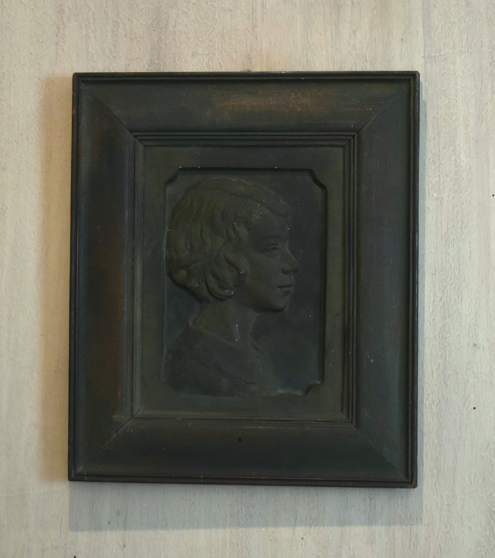 A dark bronzed plaster relief sculpture showing a profile portrait of a young person in a moulded rectangular frame.
