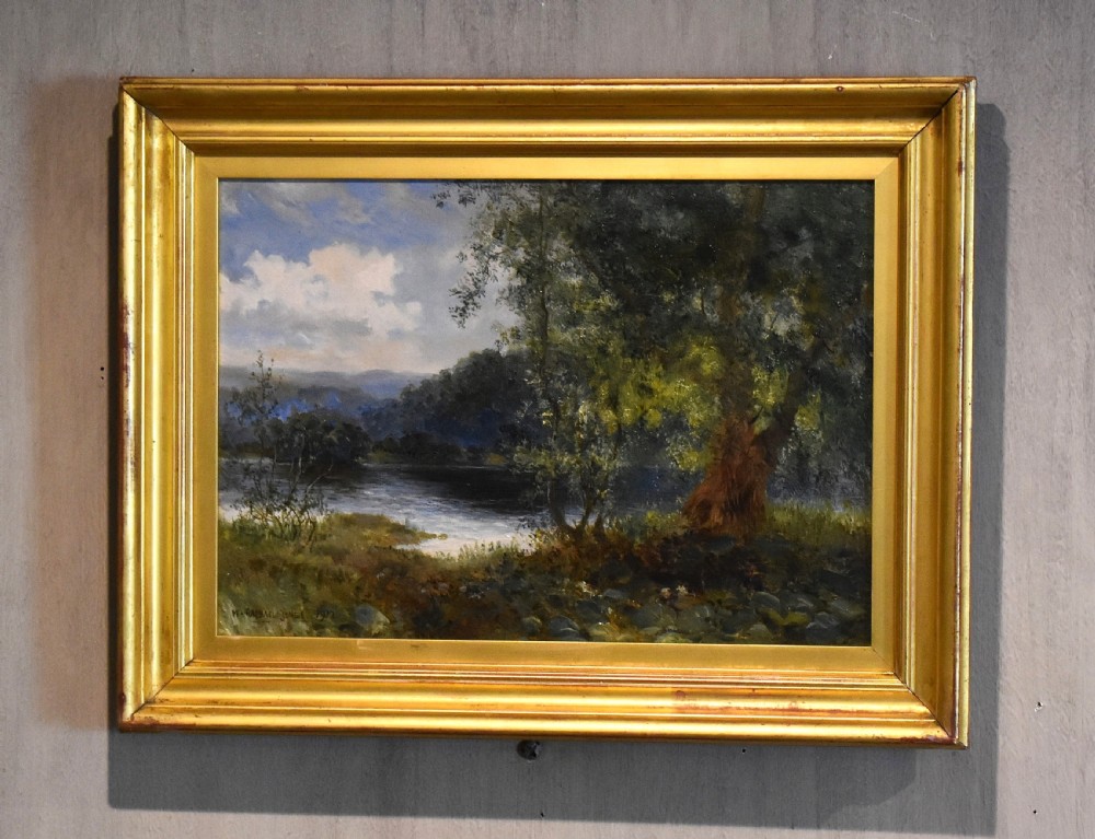 A framed oil painting depicting a tranquil river scene with leafy green trees, distant hills, and a cloudy blue sky, encased in a gold gilded wooden frame.