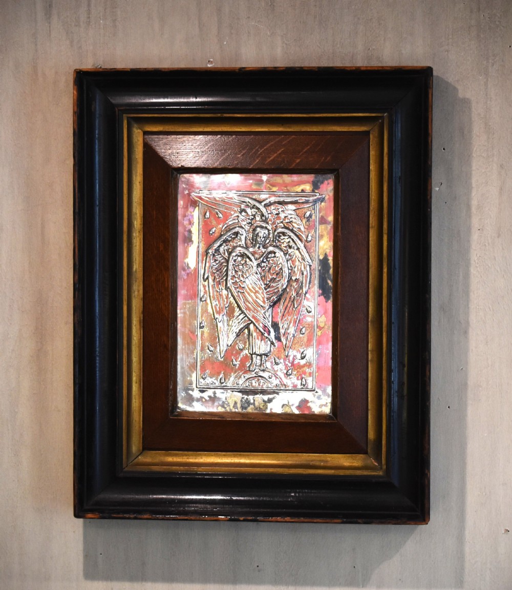An Arts and Crafts relief plaque depicting a multi-winged angel in hammered silvered metal, set within a multi-tiered ebonised and oak frame.