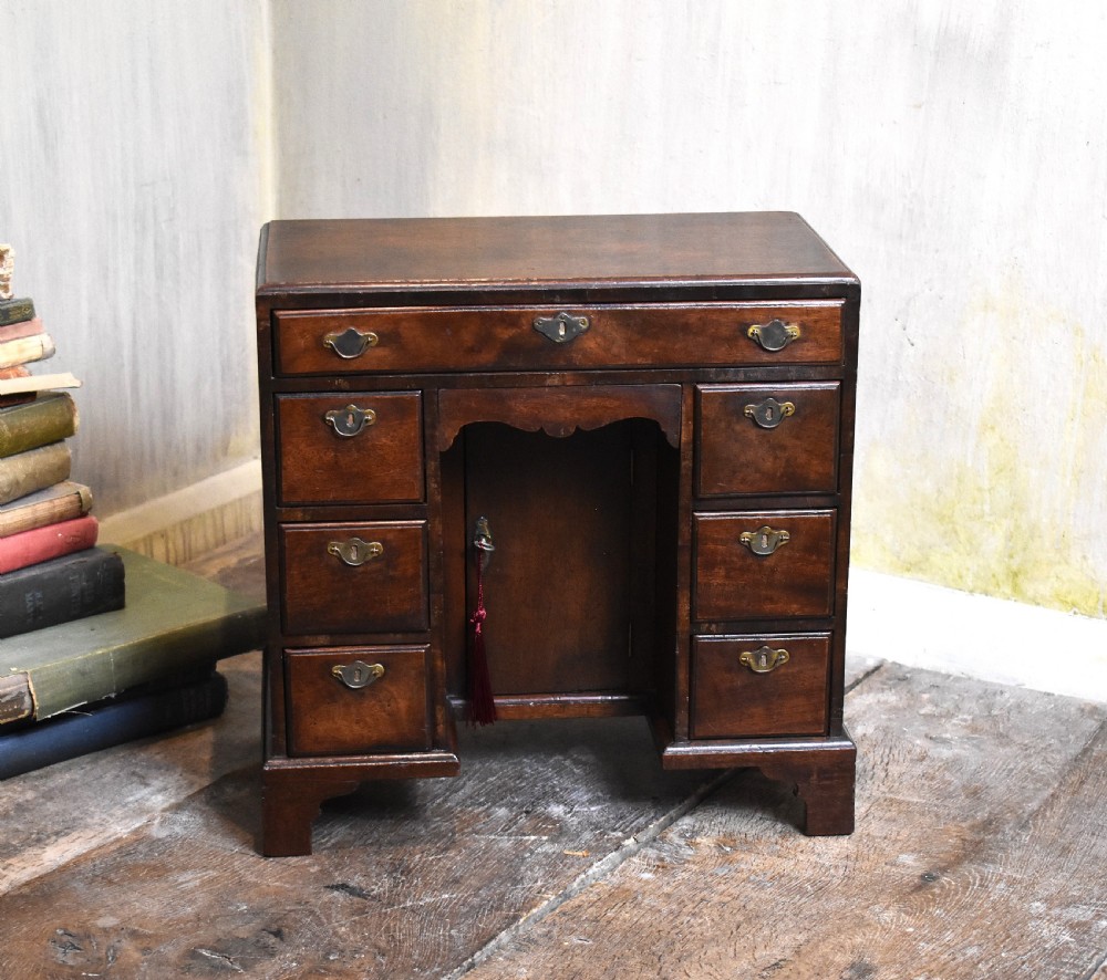 A miniature mahogany kneehole desk featuring a top drawer, six pedestal drawers, a central recessed cupboard, brass hardware, and bracket feet.