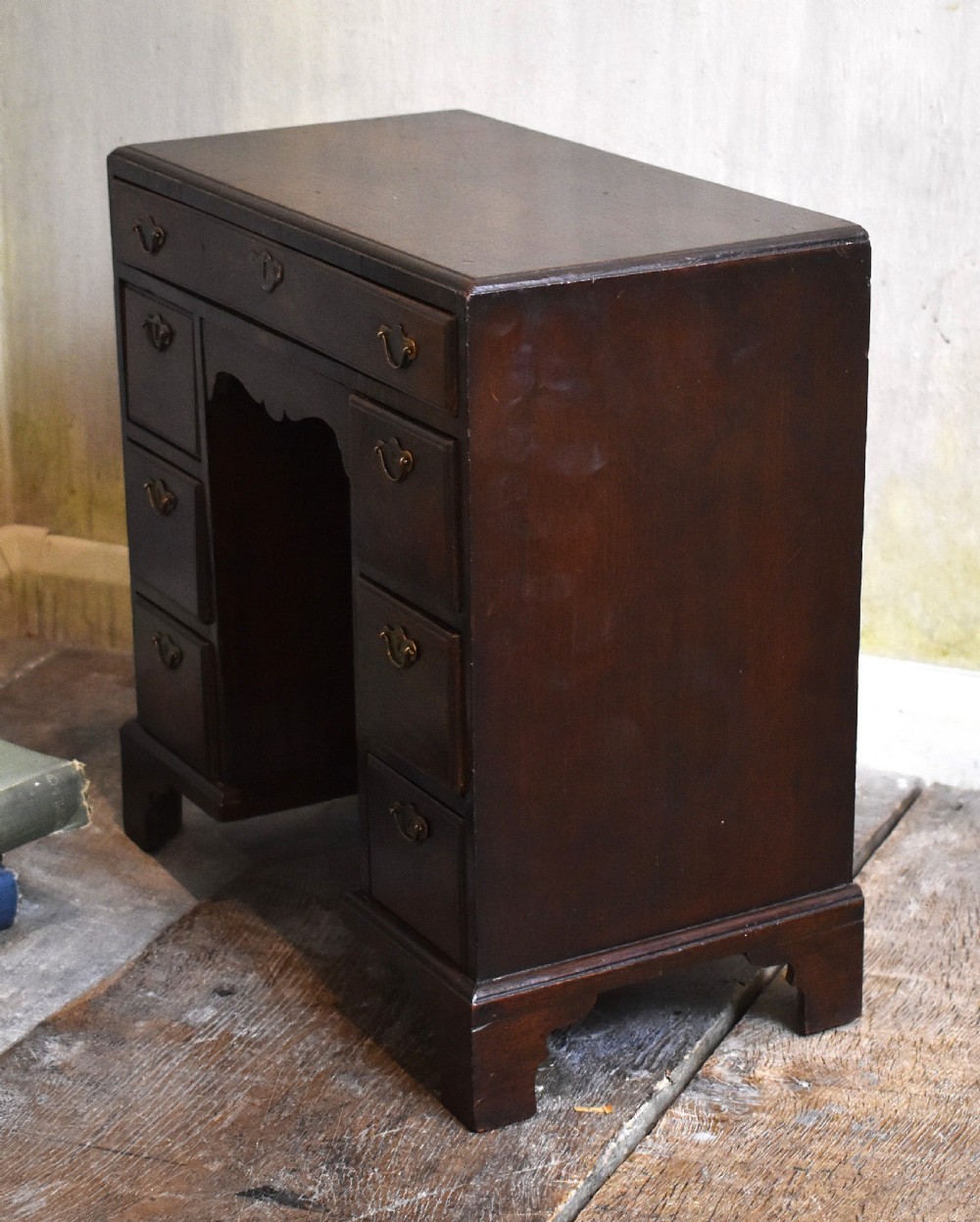 A miniature mahogany kneehole desk featuring a top drawer, six pedestal drawers, a central recessed cupboard, brass hardware, and bracket feet. View V