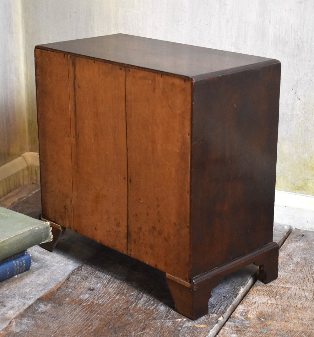 A miniature mahogany kneehole desk featuring a top drawer, six pedestal drawers, a central recessed cupboard, brass hardware, and bracket feet. View VI