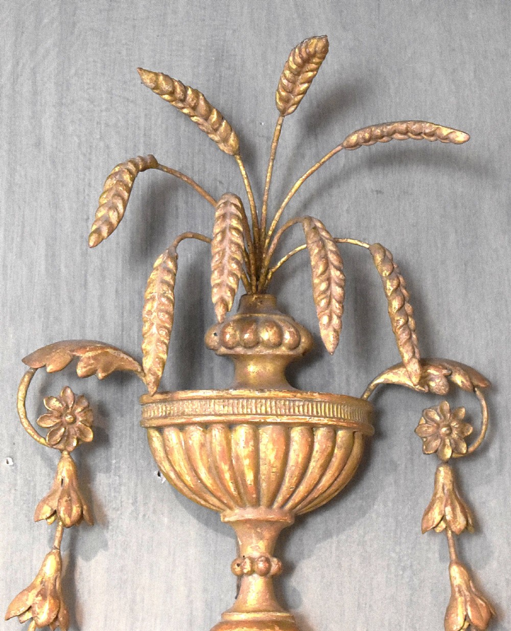 An oval Georgian giltwood mirror featuring a carved urn surmount filled with wheat sheaves, trailing floral husk swags, and an acanthus leaf bottom finial. View III